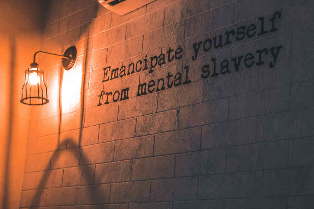Sconce attached to wall with downward bright bulb lighting wall - which is writen on with typeface saying 'Emancipate yourself from mental slavery'.